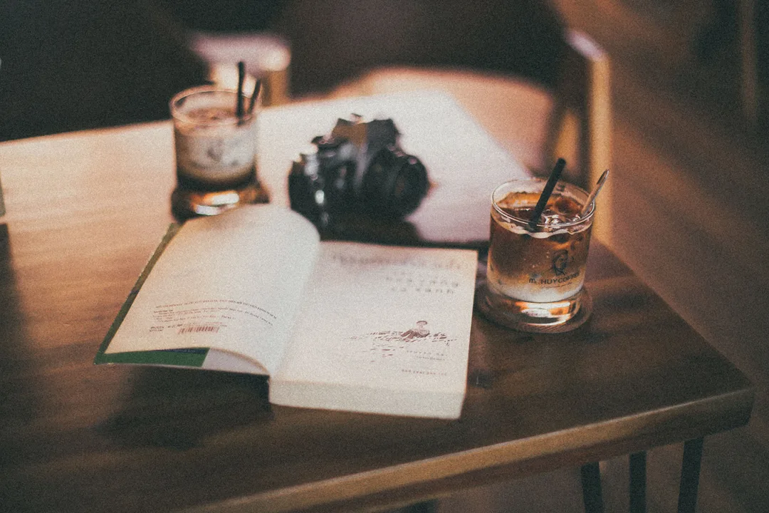 Appareil photo argentique posé sur une table en bois à côté d'un livre ouvert et de verres de café glacé